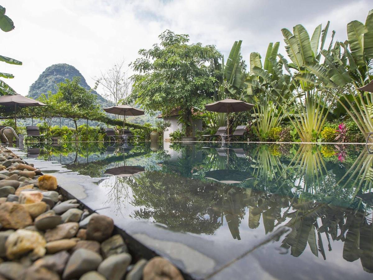 Outdoor pool edged with river stones, palms and a parasol, a karst peak rising above the garden