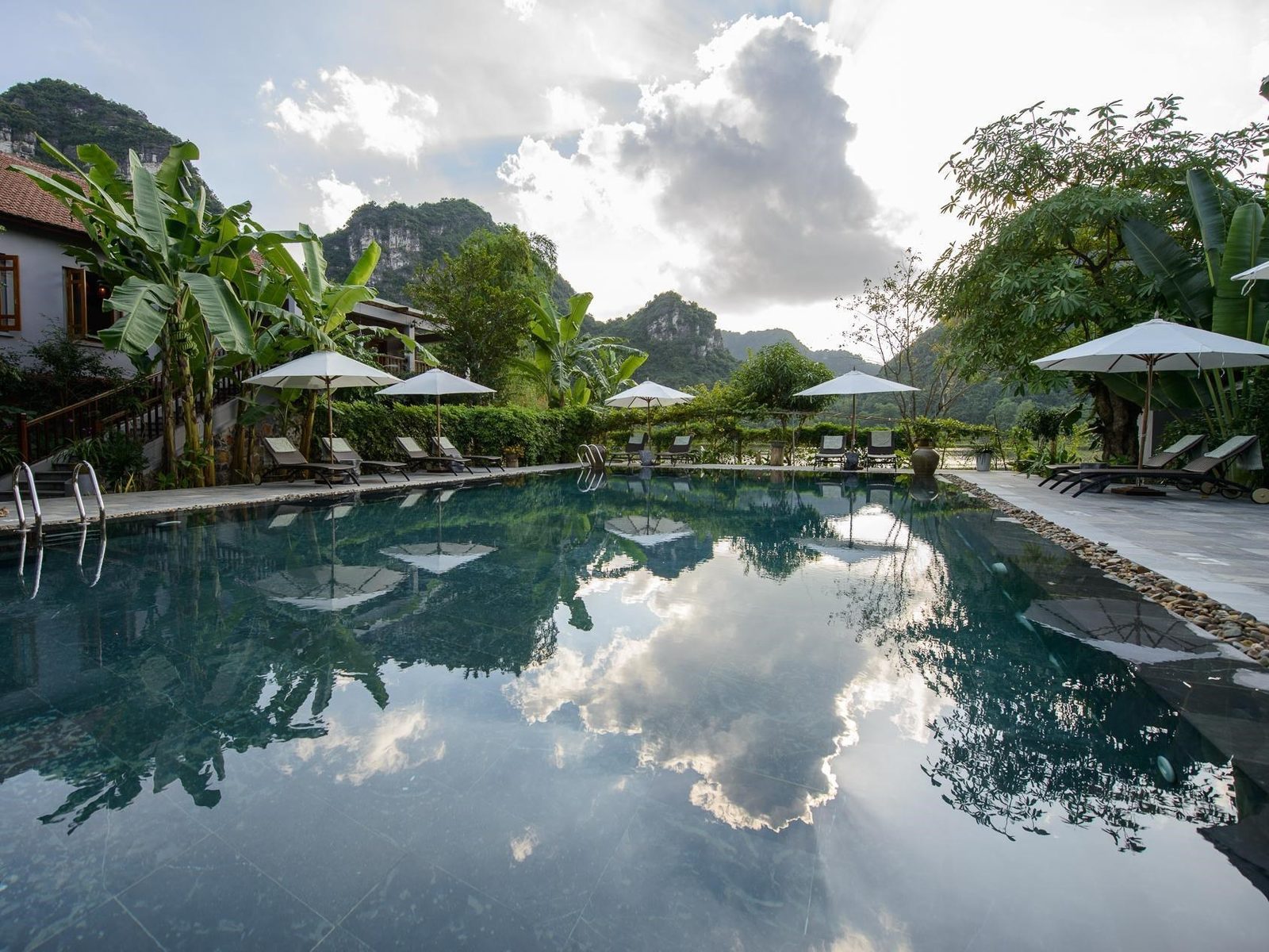 Long outdoor pool reflecting white parasols, palms and a limestone peak under a cloudy sky