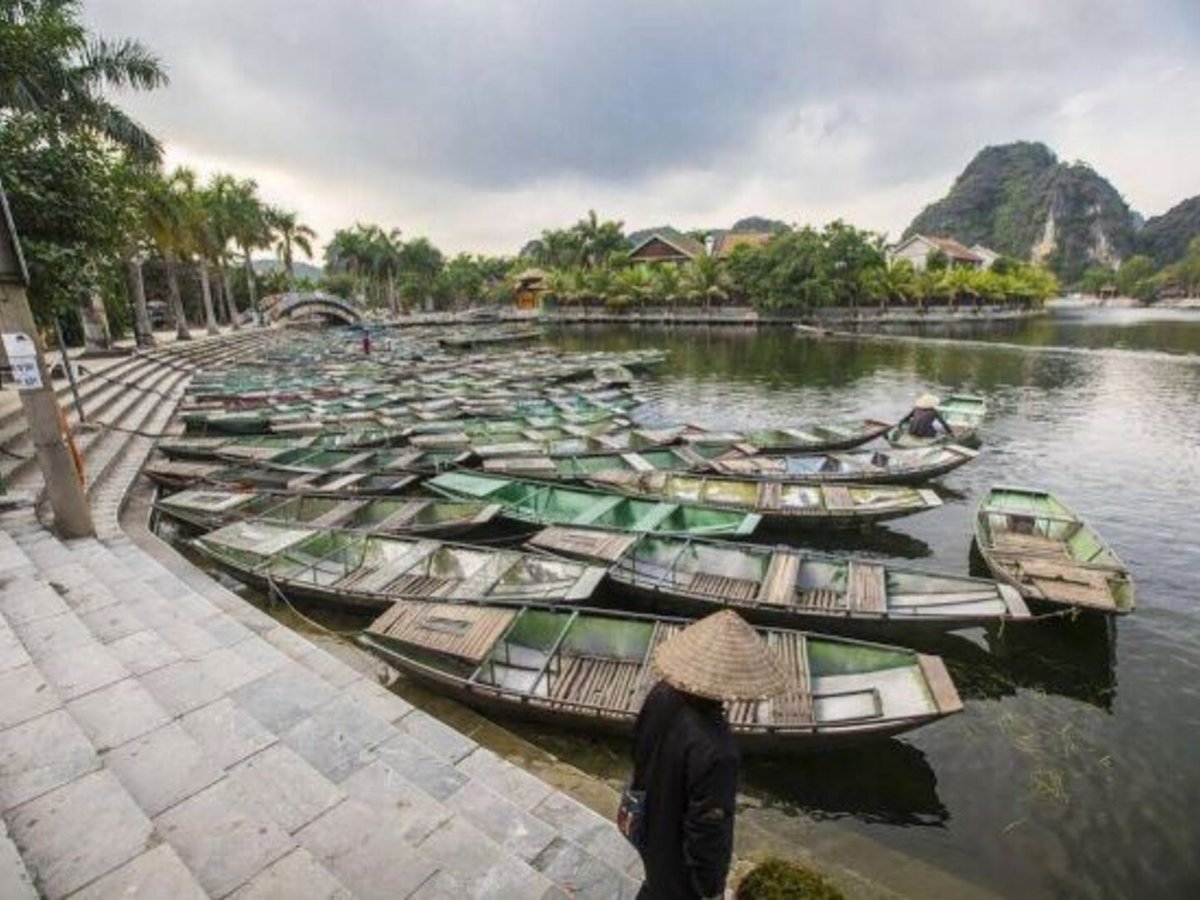 Rows of small green rowing boats tied up at a stone landing, with a limestone hill beyond the water