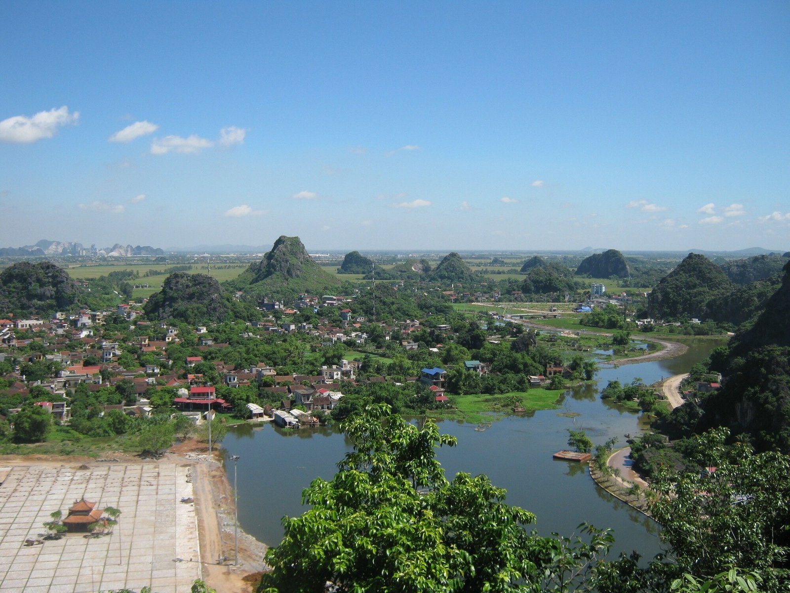 Elevated view over a village, rice fields and a winding river toward scattered limestone peaks