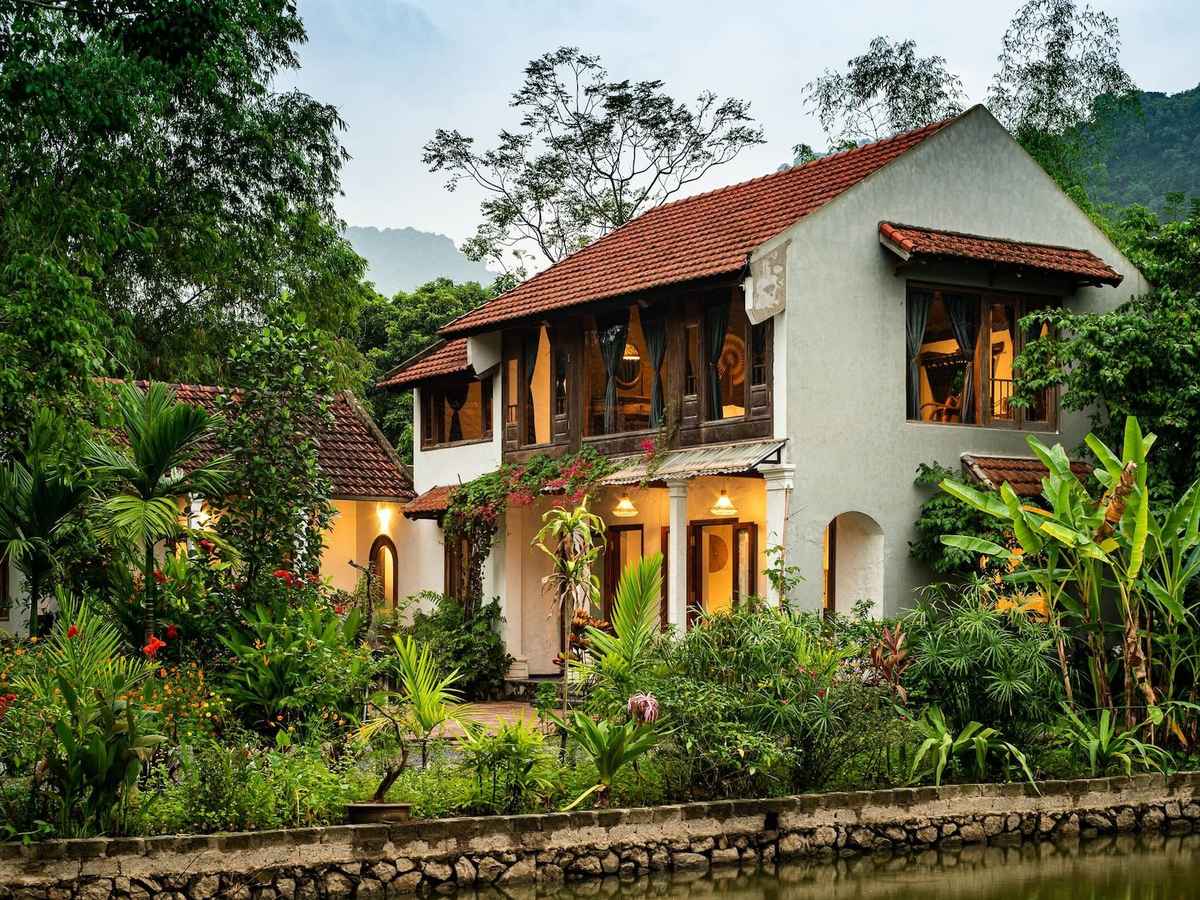 Two-story white house with a tiled roof and wooden balconies behind a garden of banana trees and flowers