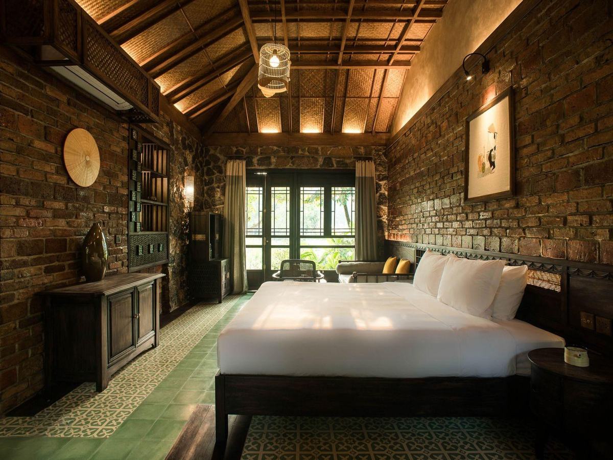 Bedroom with an exposed brick wall, dark timber ceiling, king bed and tall windows onto greenery