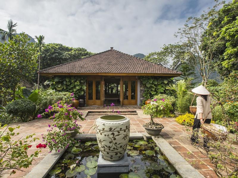 A tiled pavilion behind a lotus pond with a large glazed jar, flowering shrubs and karst hills beyond